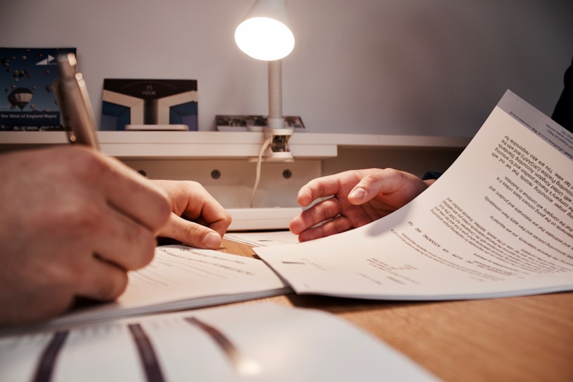 Two hands are visible, one holding a pen and another holding a document, suggesting a situation involving document review or signing. The scene appears to be set on a desk with a table lamp illuminating the area.