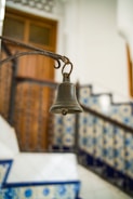 A rustic metal bell hangs from an ornate iron arm, set against a background of tiled stairs. The tiles feature intricate blue and white patterns, providing a traditional and decorative context. A wooden door is visible in the background, adding to the warm ambiance.