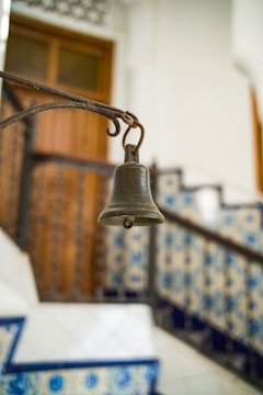 A rustic metal bell hangs from an ornate iron arm, set against a background of tiled stairs. The tiles feature intricate blue and white patterns, providing a traditional and decorative context. A wooden door is visible in the background, adding to the warm ambiance.