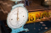 A vintage household scale with a weathered blue frame and a white dial displaying weight measurements. Behind the scale, a partially obscured yellow and orange tin with text can be seen, contributing to the nostalgic feel.