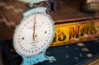 A vintage household scale with a weathered blue frame and a white dial displaying weight measurements. Behind the scale, a partially obscured yellow and orange tin with text can be seen, contributing to the nostalgic feel.