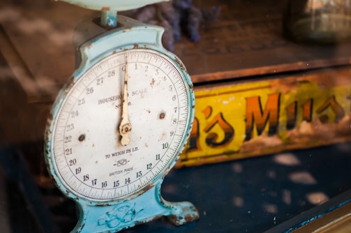 A vintage household scale with a weathered blue frame and a white dial displaying weight measurements. Behind the scale, a partially obscured yellow and orange tin with text can be seen, contributing to the nostalgic feel.