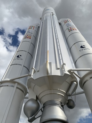A large rocket or space vehicle is positioned under a cloudy sky, with logos of ESA and CNES visible on its sides. The structure is predominantly white with some metallic components and features several flags.