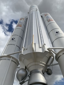 A large rocket or space vehicle is positioned under a cloudy sky, with logos of ESA and CNES visible on its sides. The structure is predominantly white with some metallic components and features several flags.