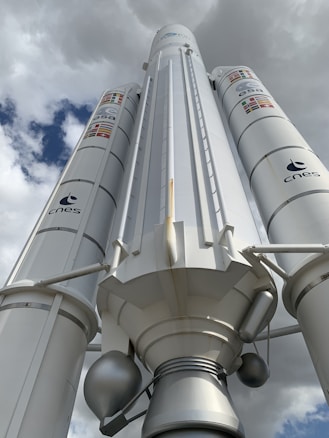 A large rocket or space vehicle is positioned under a cloudy sky, with logos of ESA and CNES visible on its sides. The structure is predominantly white with some metallic components and features several flags.
