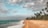 A tropical beach scene with golden sand stretching along the coast and gentle waves lapping at the shore. Tall palm trees line the edge of the beach, swaying slightly in the breeze. The sky is partly cloudy with patches of blue peeking through.
