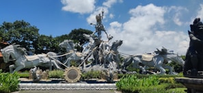 A large, intricate statue depicts a warrior figure riding a chariot pulled by multiple galloping horses. The scene is highly detailed with the warrior holding a long staff. The statue is surrounded by lush greenery and vibrant plants, set against a backdrop of a blue sky with some clouds.