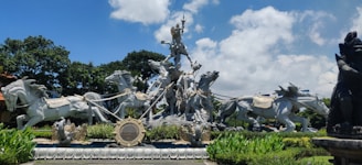 A large, intricate statue depicts a warrior figure riding a chariot pulled by multiple galloping horses. The scene is highly detailed with the warrior holding a long staff. The statue is surrounded by lush greenery and vibrant plants, set against a backdrop of a blue sky with some clouds.