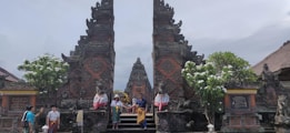 A traditional Balinese temple with intricate carvings is flanked by tall, ornately decorated gates. Several people in colorful attire are sitting and standing around the entrance. Lush greenery and flowering trees are present, adding vibrant color to the historic scene.