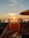 A hand holds a frosty glass with a straw, filled with a brightly colored fruity drink. The background reveals a scenic sunset view over the ocean, with palm trees silhouetted against the colorful sky. An umbrella provides shade on a patio or deck area overlooking the beach.