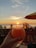 A hand holds a frosty glass with a straw, filled with a brightly colored fruity drink. The background reveals a scenic sunset view over the ocean, with palm trees silhouetted against the colorful sky. An umbrella provides shade on a patio or deck area overlooking the beach.
