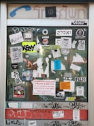 A glass door is covered with a variety of stickers and graffiti. The stickers include logos, text in multiple languages, and symbols. Some notable elements are the 'ANTIFA DRESDEN' sticker, the 'NETFLIX and chill' sticker, and a warning sign in multiple languages about a 5,000 Baht fine for advertising in the area. The door frame has a vintage design with faded blue and red signage.