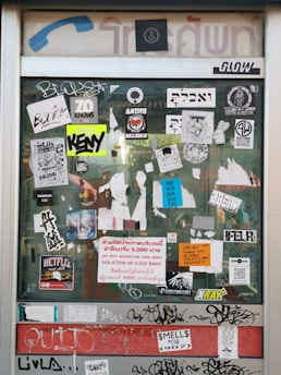 A glass door is covered with a variety of stickers and graffiti. The stickers include logos, text in multiple languages, and symbols. Some notable elements are the 'ANTIFA DRESDEN' sticker, the 'NETFLIX and chill' sticker, and a warning sign in multiple languages about a 5,000 Baht fine for advertising in the area. The door frame has a vintage design with faded blue and red signage.