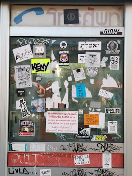 A glass door is covered with a variety of stickers and graffiti. The stickers include logos, text in multiple languages, and symbols. Some notable elements are the 'ANTIFA DRESDEN' sticker, the 'NETFLIX and chill' sticker, and a warning sign in multiple languages about a 5,000 Baht fine for advertising in the area. The door frame has a vintage design with faded blue and red signage.