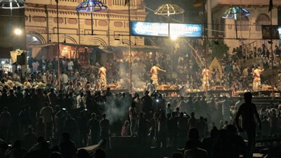 A large gathering of people is attending a cultural or religious event at night. Several individuals are dressed in traditional clothing, performing rituals on a stage. The background features a historic building with intricate architecture, illuminated by artificial lights. Crowds of spectators are visible in the dimly lit surroundings, creating an atmospheric scene.