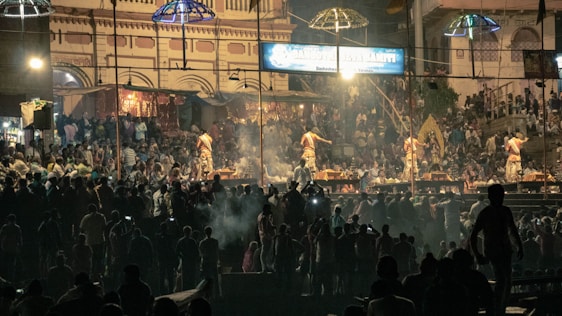 A large gathering of people is attending a cultural or religious event at night. Several individuals are dressed in traditional clothing, performing rituals on a stage. The background features a historic building with intricate architecture, illuminated by artificial lights. Crowds of spectators are visible in the dimly lit surroundings, creating an atmospheric scene.