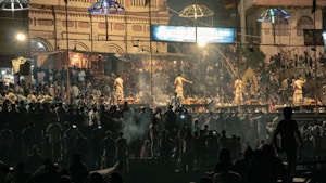 A large gathering of people is attending a cultural or religious event at night. Several individuals are dressed in traditional clothing, performing rituals on a stage. The background features a historic building with intricate architecture, illuminated by artificial lights. Crowds of spectators are visible in the dimly lit surroundings, creating an atmospheric scene.