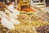 Various types of olives and possibly other preserved foods are displayed in a market setting. Large white scoops are placed over the piles of food items, allowing customers to serve themselves. The setup suggests a vibrant, bustling market atmosphere.