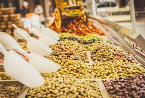 Various types of olives and possibly other preserved foods are displayed in a market setting. Large white scoops are placed over the piles of food items, allowing customers to serve themselves. The setup suggests a vibrant, bustling market atmosphere.