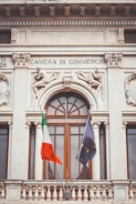 A historic building facade with ornate sculptures and decorative elements, featuring two flags in the foreground. The architecture has classical design elements, including columns and intricate stone carvings. Above a large window, the text 'Camera di Commercio' is inscribed.