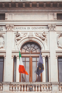 A historic building facade with ornate sculptures and decorative elements, featuring two flags in the foreground. The architecture has classical design elements, including columns and intricate stone carvings. Above a large window, the text 'Camera di Commercio' is inscribed.