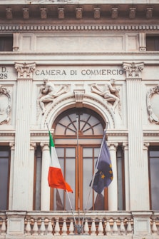 A historic building facade with ornate sculptures and decorative elements, featuring two flags in the foreground. The architecture has classical design elements, including columns and intricate stone carvings. Above a large window, the text 'Camera di Commercio' is inscribed.