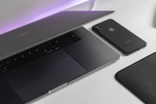 A sleek, modern workspace featuring a partially opened laptop alongside a smartphone. Both devices have a minimalist design with a focus on clean lines and a muted color palette. The background is softly illuminated with a subtle purple light, adding a contemporary touch.