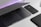 A sleek, modern workspace featuring a partially opened laptop alongside a smartphone. Both devices have a minimalist design with a focus on clean lines and a muted color palette. The background is softly illuminated with a subtle purple light, adding a contemporary touch.