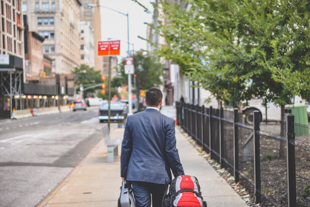 person in black suit jacket with r ed bag walking beside metal fence