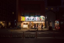 A noodle shop is illuminated at night with warm lighting, showcasing a large, colorful sign featuring images of ramen. Several people are walking past the shop, and there is a red vehicle parked on the street nearby. The surrounding area is dimly lit, creating a contrast with the bright storefront.