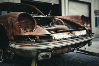 A rusted, vintage car with missing components positioned in front of a garage with open doors. The car appears significantly deteriorated, with visible corrosion and missing headlights, emphasizing its neglected state.