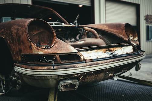 A rusted, vintage car with missing components positioned in front of a garage with open doors. The car appears significantly deteriorated, with visible corrosion and missing headlights, emphasizing its neglected state.