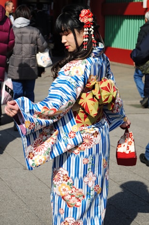 A woman dressed in a traditional Japanese kimono stands among a group of people. The kimono features blue and white stripes with floral patterns, and she is holding a matching handbag. Her hair is adorned with red accessories.