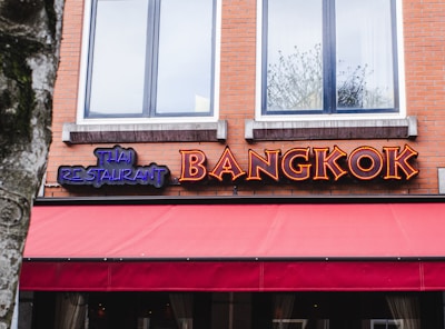 A restaurant storefront with large, vibrant neon signs that spell out 'THAI RESTAURANT' in blue and 'BANGKOK' in orange against a brick wall. The restaurant features two upper windows and a red awning.