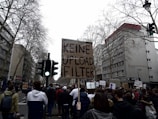 A large group of people is gathered on a city street, participating in a protest. The central focus is a sign held high, reading 'KEINE UPLOAD FILTER' in bold letters. The street is lined with tall, bare trees and multi-story buildings. A traffic light is visible with a green light, and the sky appears overcast.