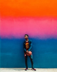 A young person wearing casual sports clothing holds a basketball while standing against a brightly colored wall. The wall features a gradient that transitions from orange to pink to blue.