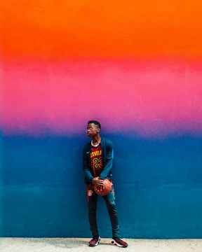 A young person wearing casual sports clothing holds a basketball while standing against a brightly colored wall. The wall features a gradient that transitions from orange to pink to blue.