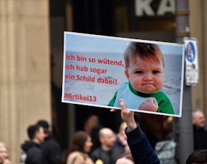 A hand holding a protest sign with an image of a determined-looking baby clenching a fist. The sign includes German text related to Article 13. There are people and a building in the blurred background.