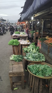 A bustling street market with numerous stalls displaying a variety of fresh vegetables including lettuce, cabbage, and other leafy greens. Vendors are present, and the atmosphere is lively with people walking by. The market is set under large tarps, providing shade and some lighting is visible, giving a cozy yet vibrant ambiance.