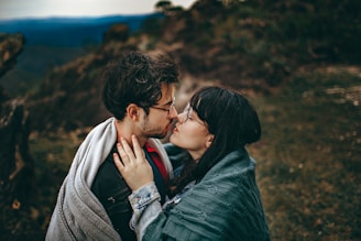 A couple stands closely together wrapped in cozy blankets, sharing an intimate moment against a blurry natural backdrop. The man has a beard and wears glasses, while the woman has long dark hair and also wears glasses.