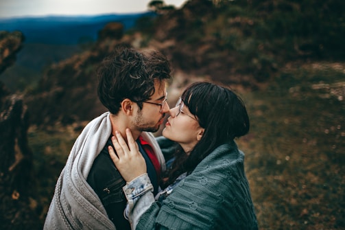 A couple stands closely together wrapped in cozy blankets, sharing an intimate moment against a blurry natural backdrop. The man has a beard and wears glasses, while the woman has long dark hair and also wears glasses.