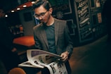A man with glasses reads a magazine in a dimly lit, stylish cafe. He is wearing a tweed blazer and has neatly styled hair. The background features dark, chalkboard-style artwork and warm lighting from overhead.