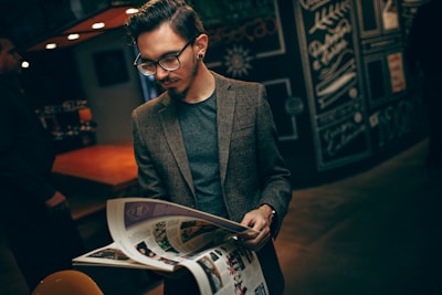 A man with glasses reads a magazine in a dimly lit, stylish cafe. He is wearing a tweed blazer and has neatly styled hair. The background features dark, chalkboard-style artwork and warm lighting from overhead.