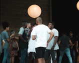 A group of people are gathered casually under soft lighting. They appear to be in conversation and socializing, with one person laughing. A large paper lantern emits warm light, creating a cozy atmosphere. The scene suggests a relaxed setting.