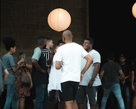 A group of people are gathered casually under soft lighting. They appear to be in conversation and socializing, with one person laughing. A large paper lantern emits warm light, creating a cozy atmosphere. The scene suggests a relaxed setting.