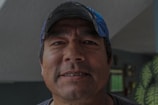 A middle-aged man with a rugged complexion is smiling slightly. He is wearing a blue baseball cap and a dark-colored shirt, and stands indoors with a wall in the background that has some decorations and a window.