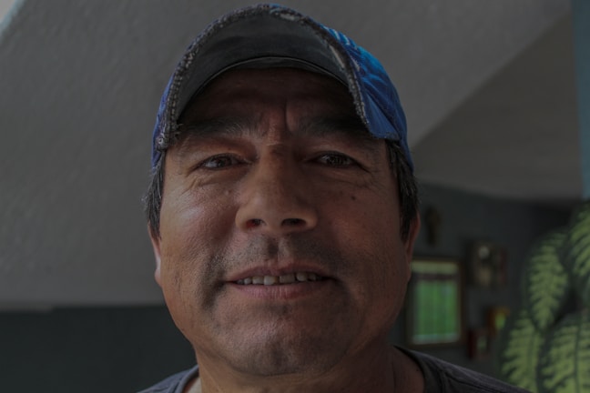 A middle-aged man with a rugged complexion is smiling slightly. He is wearing a blue baseball cap and a dark-colored shirt, and stands indoors with a wall in the background that has some decorations and a window.