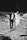 A man and a woman dressed in formal attire, holding hands while walking on a rocky shoreline. The woman is wearing a long white dress and the man is wearing a dark suit. The man is carrying a bouquet of flowers in one hand.