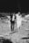A man and a woman dressed in formal attire, holding hands while walking on a rocky shoreline. The woman is wearing a long white dress and the man is wearing a dark suit. The man is carrying a bouquet of flowers in one hand.