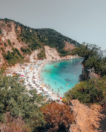 A scenic coastal area featuring a secluded beach with numerous white parasols and sunbathers enjoying the turquoise waters. The backdrop consists of rugged cliffs and lush greenery, providing a dramatic and natural setting.